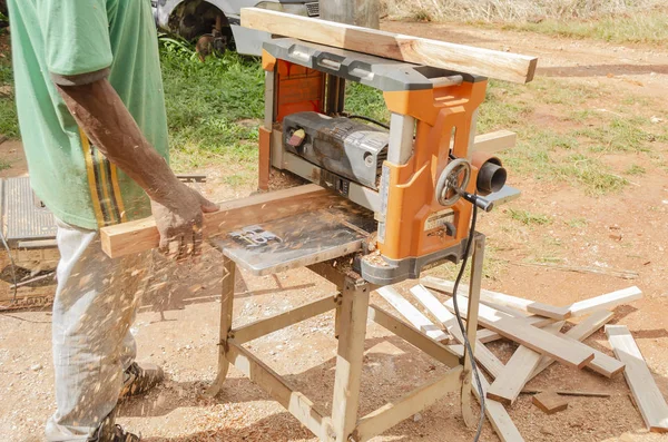 A cabinet maker guiding a piece of wood along the table and through an electric planer that is seated on a wooden stand. As the wood passes through the machine, saw dust propels in the opposite direction the wood moves, covering the man`s hand.
