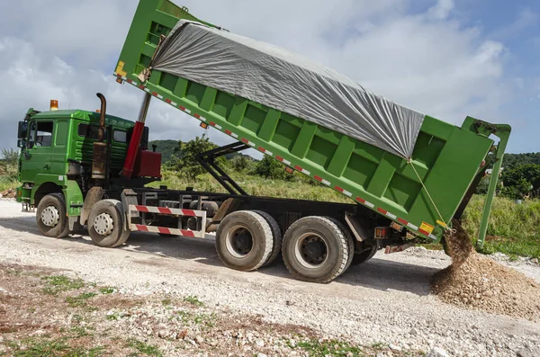 Looking sideways on a green tipper truck with its body covered with grey tarpaulin as it gradually rises to empty small amounts of its marl content on a road that is in the process of being constructed.