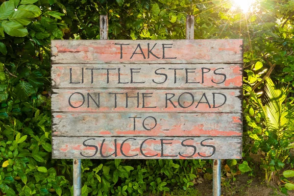Kleine Schritte auf dem Weg zum Erfolg Motivationszitat auf altem Vintage-Schild im Wald geschrieben, mit Sonnenstrahlen im Hintergrund. — Stockfoto