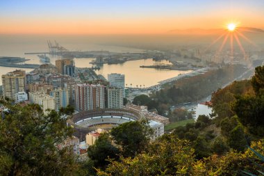 Malaga 'da günbatımı. İspanyol Costa del Sol şehrinin akşam havasında limanın panoramik manzarası, evler, ağaçlar, aydınlatılmış sokak lambası ve dağların üzerinde mavi gökyüzü