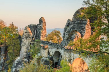 Saxon İsviçre 'deki Bastei Köprüsü. Ulusal Saksonya parkındaki tarihi binalar. Sonbahar havasında akşamları rock yapmak. Ağaçların yeşil, kahverengi ve sarı yaprakları