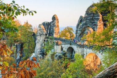 Saxon İsviçre 'deki Bastei Köprüsü. Unesco Ulusal Saksonya Parkı 'ndaki tarihi binalar. Sonbahar havasında akşam vakti patikalı rock. Ağaçların yeşil, kahverengi ve sarı yaprakları