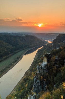 Akşamları Elbe Vadisi 'nde Sakson İsviçre. Bastei Köprüsü manzaralı ulusal park Elbe ve kayalar, ağaçlar ve ormanlar sonbahar havasında ve gün batımında turuncu bir ufuk ile