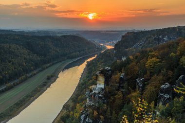 Akşamları Elbe Vadisi 'nde Sakson İsviçre. Bastei Köprüsü 'nden Elbe' ye uzanan milli parktaki manzara ve sonbahar havasında kayalar, ağaçlar ve ormanlar gün batımında turuncu bir ufuk ile birlikte.