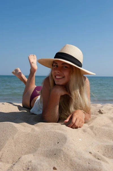 The girl in the hat lies on the beach near the sea.