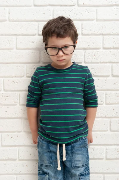 Niño sombrío en gafas se levanta sobre un fondo de una pared de ladrillo blanco — Foto de Stock