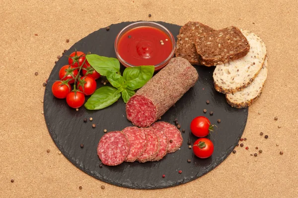 Gesneden rookworst met saus, kerstomaten en basilicum op een zwart grafietbord met sneetjes brood. bovenaanzicht — Stockfoto