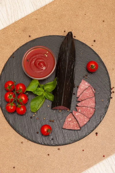 Krájená uzená klobása s omáčkou, cherry rajčaty a bazalkou na černé grafitové desce. pohled shora — Stock fotografie