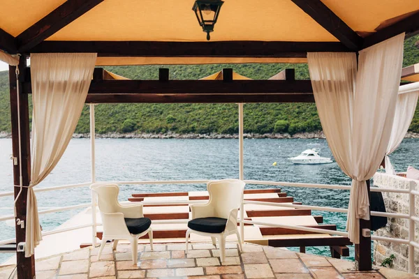 Duas cadeiras brancas com travesseiros pretos ficam em um gazebo junto ao mar com vista para um barco branco, em Montenegro, na Península Lústica — Fotografia de Stock