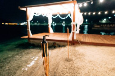 Deniz kıyısında meşale. Gece kumsalda parti var. Çelenkli bir pergola.