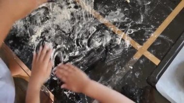 Bakıcı annelerin elleri hamur yoğurur, çocukların elleri yardım eder. Siyah masa ve beyaz un. Çocuklar yemek yapmakla ilgilenirler. Çocuklar masada beyaz buğday unuyla oynuyorlar..