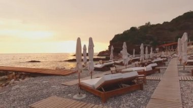 Rocky Sahili 'ndeki çift güneşli yatakların üzerinde kamera uçurmak. Yumuşak şilteli ve beyaz şemsiyeli iki kişilik yatak. Çakıl taşı plajında ahşap bir patika. Karadağ 'da Adriyatik Kıyısı