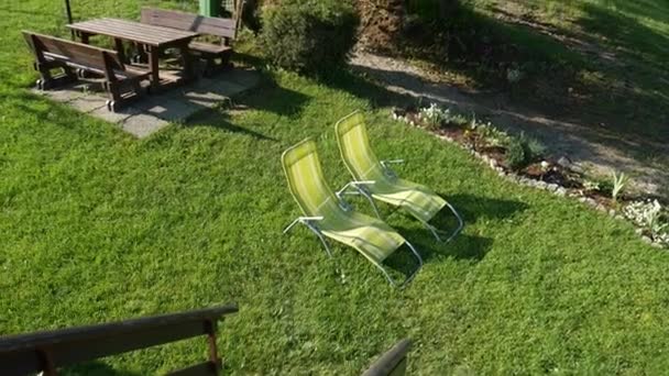 Metalen ligstoelen in de tuin op een groen gazon en een houten tafel en banken. Een prachtige goed verzorgde tuin met een recreatiegebied en eetgelegenheid. — Stockvideo