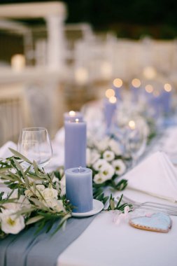 Resepsiyondaki düğün yemeği masasının yakın çekimi. Mavi, kalın mumlar masada beyaz çiçeklerin ve zeytin dallarının arasında yanar.