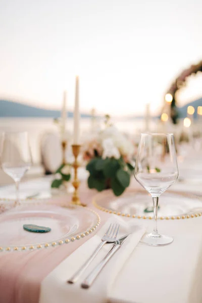 Close-up van de bruiloft eettafel. Prachtig glaswerk - borden met gouden kralen, wijnglazen, messen en vorken, op achtergrond van kaarsen en bloemstukken. Bruiloft buiten, zonsondergang achtergrond — Stockfoto