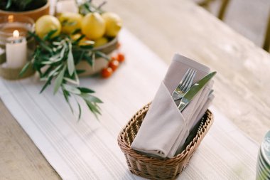 Bıçak ve çatal yemek odası peçetesine doldurulmuş hasır sepetteki ahşap bir masada. Arka planda zeytin dalı, limon, kiraz domatesli bir tabak ve şamdanda bir mum yanıyor.