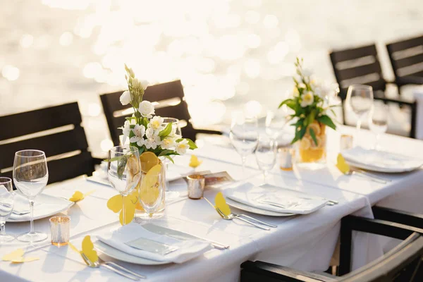 Bruiloft diner tafel receptie. Rechthoekige tafels met wit tafelkleed, bloemstukken citroenen in vazen. Gele papieren vlinders liggen verspreid op tafel. Op het strand, met uitzicht op zonsondergang op zee. — Stockfoto