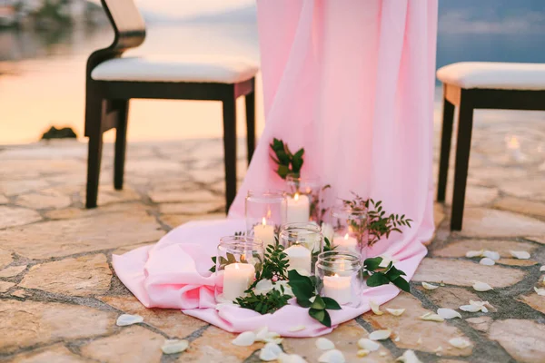 Primo piano di un tavolo da pranzo nuziale alla reception. Molte candele accese stanno ai piedi del tavolo su una tovaglia di seta rossa, i petali di rosa sono sparsi su piastrelle di pietra, il tavolo si erge al tramonto. . — Foto Stock