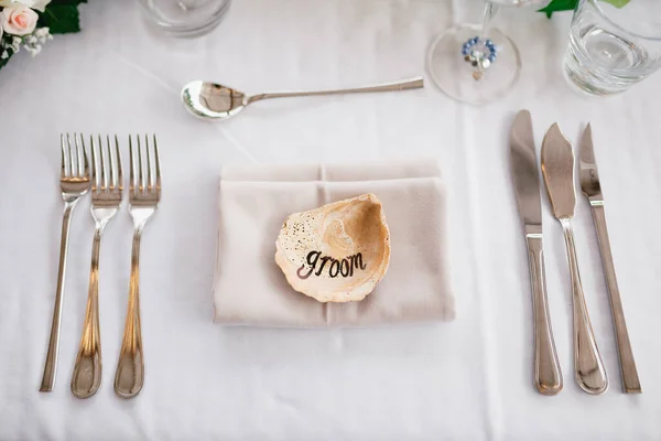 Close-up of a wedding dinner table. The inscription GROOM on the shell in center of the table. Serving a table - knives, forks, glasses, wine glasses, against background of gray tablecloth