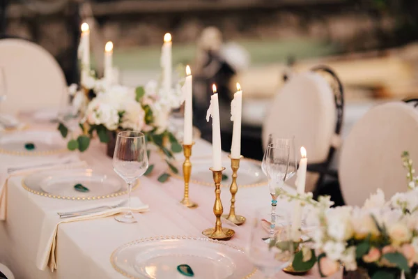 Wedding dinner table at reception. Beautiful white delicate candles burn in metal candlesticks, against background of white and pink tablecloth, flower arrangements and glass plates with gold beads — Stock Photo, Image