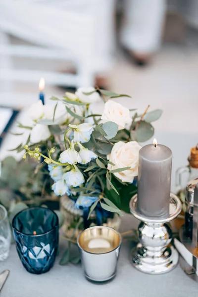 Zbliżenie weselnego stołu w recepcji. Duża gruba szara świeca stoi w srebrnym błyszczącym świeczniku, obok kompozycji kwiatowej i z małą tabliczką świec w niebieskiej szklance. — Zdjęcie stockowe