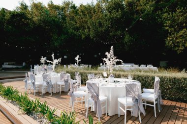 Yuvarlak masalar, beyaz kumaş masa örtüsü, beyaz Chiavari sandalyeleri, kumaştan gri kumaşlı ve fiyonklu. Masanın ortasında mumlarla süslenmiş kristal süslemeler var. Masanın üzerinde çelenkler yanıyor..