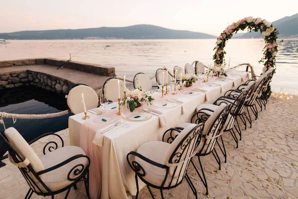 Close-up van een huwelijksreceptie. Een tafel staat op het strand met uitzicht op de bergen bij zonsondergang. Metalen gesmeed stoelen, brandende kaarsen, bruiloft boog van bloemen, room gekleurde doek tafelkleed. — Stockfoto