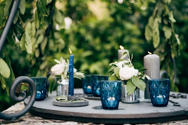 Detailní záběr svatební večeře na recepci. Šedé silné svíčky, modrá tenká svíčka, malé tabletu svíčky v modrých brýlích hořet stojící na kovovém stole na zeleném pozadí. — Stock fotografie