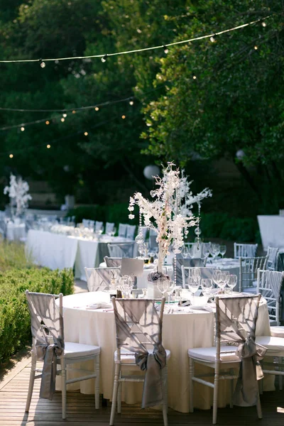 Round tables with a white fabric tablecloth, white Chiavari chairs with drapery gray fabric with a bow. In the center of the table is crystal decoration with candles, garlands are lit above the table.