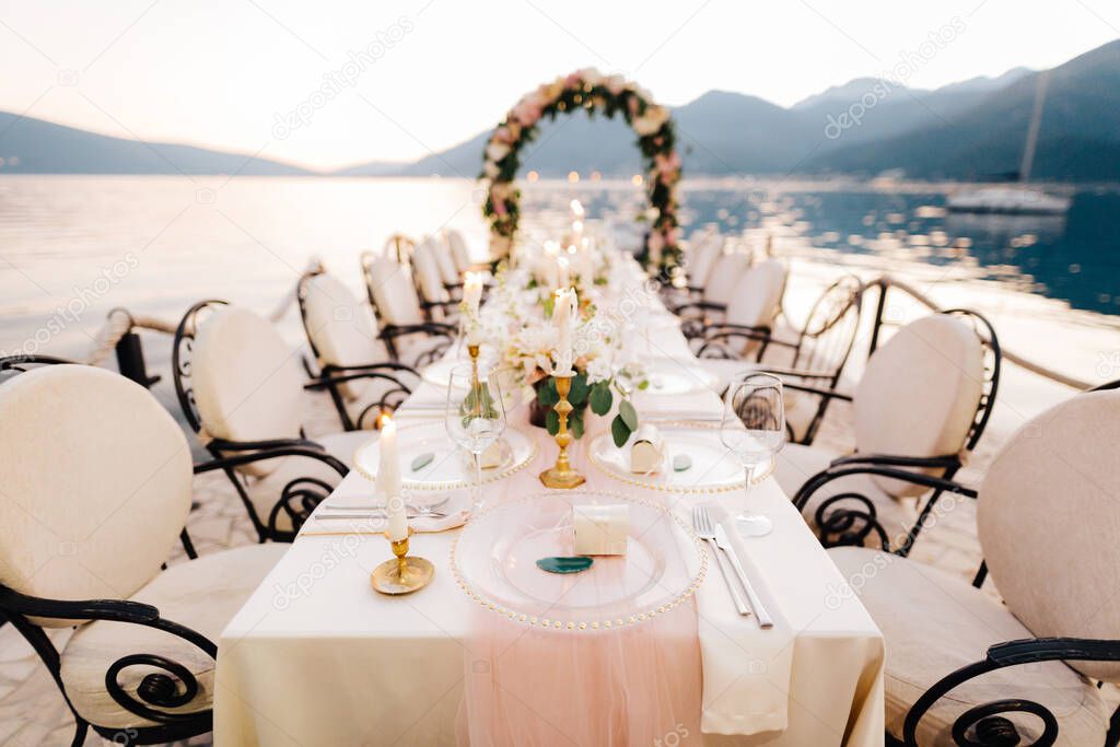 Close-up of a wedding dinner table reception. A table stands on beach overlooking mountains at sunset. Metal forged chairs, burning candles, wedding arch of flowers, cream-colored cloth tablecloth.