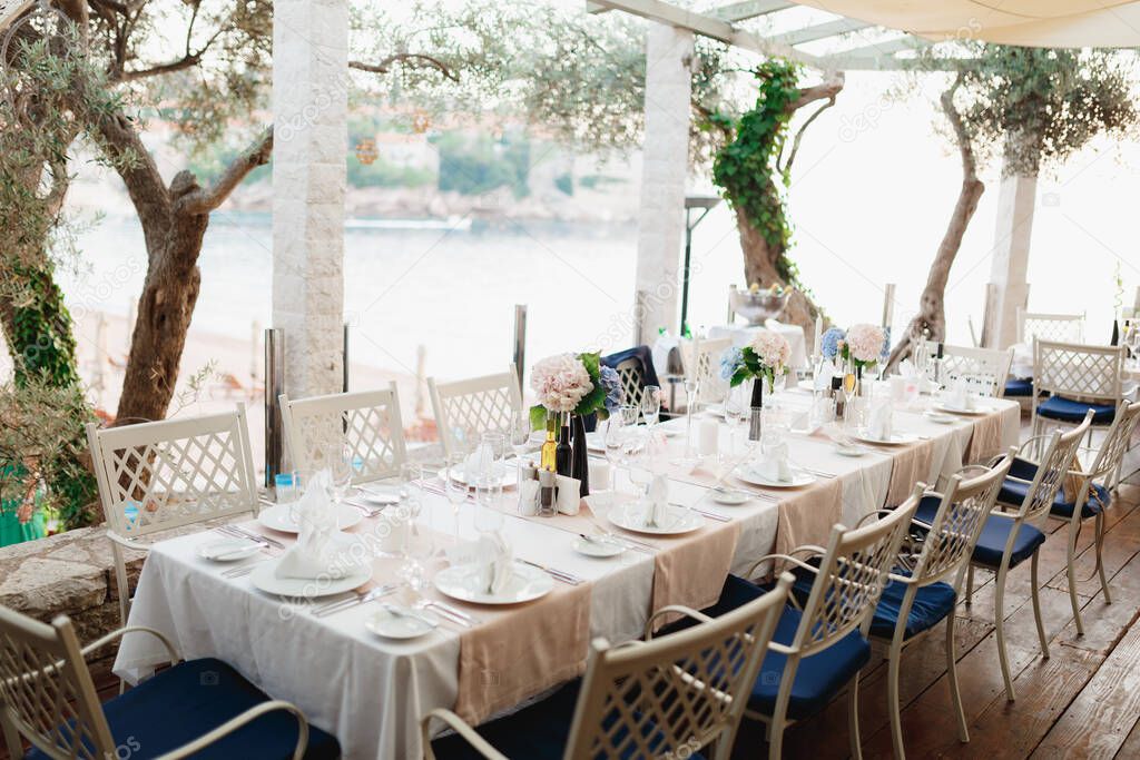 Close-up of a wedding dinner table at reception. A long table with metal chairs with blue pillows, in a stone gazebo between olive trees, overlooking Sveti Stefan Island, in Montenegro.