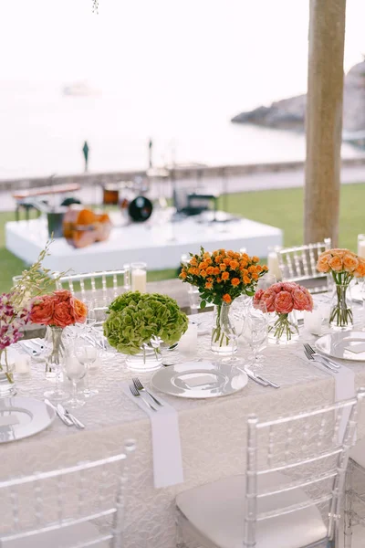 Recepción de mesa de cena de boda. Una mesa muy larga para invitados con un mantel blanco, arreglos florales, sillas transparentes de plástico de vidrio Chiavari. Bajo las viejas columnas con vides de glicina . — Foto de Stock