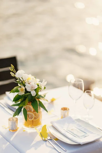 Recepción de mesa de cena de boda. Baño transparente de vidrio con limones en rodajas y un ramo de Alstromeri blanco, sobre la mesa sobre el fondo del resplandor del agua a la luz del atardecer — Foto de Stock