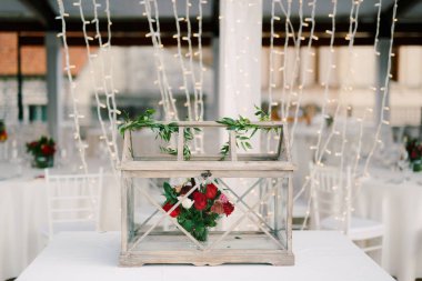 Wedding dinner table reception. Decoration of the table - a wooden box with glass, with a bouquet of flowers inside of red roses and carnations against the background of the New Years garland.  clipart