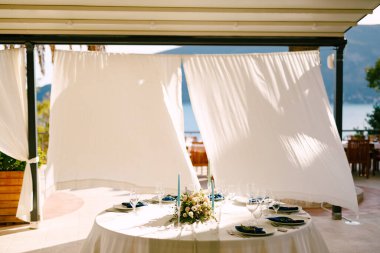 Düğün masası resepsiyonu. Ortasında bir buket çiçek olan yuvarlak masa ve rüzgarda gelişen beyaz masa örtülerinin arka planına karşı mumlar.