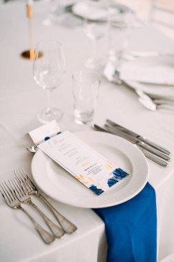 Düğün masası resepsiyonu. Masada beyaz tabak, solda üç çatal, sağda üç bıçak. Düğün menüsü tabakta. Üst görünüm