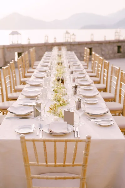 Perspectiva boda cena mesa recepción vista. Una larga mesa rectangular con un mantel crema y flores de orquídea en el centro. Contra el telón de fondo de las montañas, el mar y el atardecer — Foto de Stock