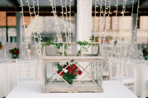 Wedding dinner table reception. Decoration of the table - a wooden box with glass, with a bouquet of flowers inside of red roses and carnations against the background of the New Years garland. — Stock Photo, Image
