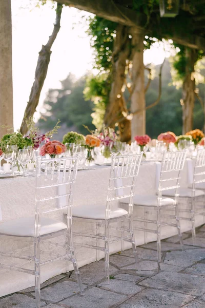 Recepción de mesa de cena de boda. Sillas de cristal transparente Chiavari, Tiffany, de pie cerca de la mesa con flores. Sobre el fondo de la luz del atardecer a través del follaje y la mesa de glicinas — Foto de Stock