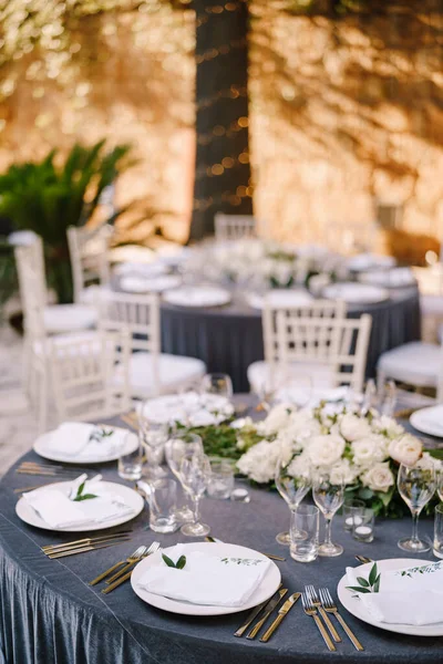 Hochzeitstisch-Empfang. Runde Banketttische für Gäste mit grauer Tischdecke. Blumengesteck aus Rosen in der Mitte des Tisches. Girlanden an Baumstämmen, Kristallleuchter hängen in der Luft — Stockfoto