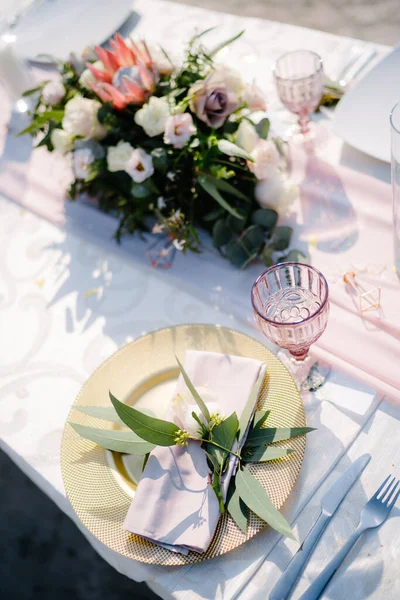 Прием свадебного ужина. Золотая тарелка с розовой салфеткой и оливковыми листьями. Старое малиновое стекло. Цветочная композиция в центре стола . — стоковое фото