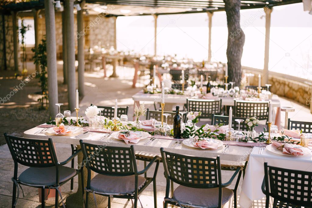 Wedding dinner table reception. There are many rectangular tables for guests with a pink tablecloth, in the rays of the sunset on the beach. Heavy metal chairs with a mesh back. 