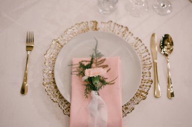 Düğün masası resepsiyonu. Beyaz, pembe kumaş havlunun altında altın tabak, üstünde ipek kurdele olan pembe çiçek. Tabağın yanındaki çatal bıçak takımı. Üst görünüm