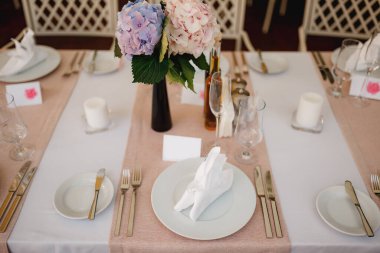 Düğün masası resepsiyonu. Masadaki tabakların yakın çekimi, çatal bıçak takımı, şarap bardakları, şamdanlardaki mumlar. Masada bir buket mavi ve pembe ortanca var..