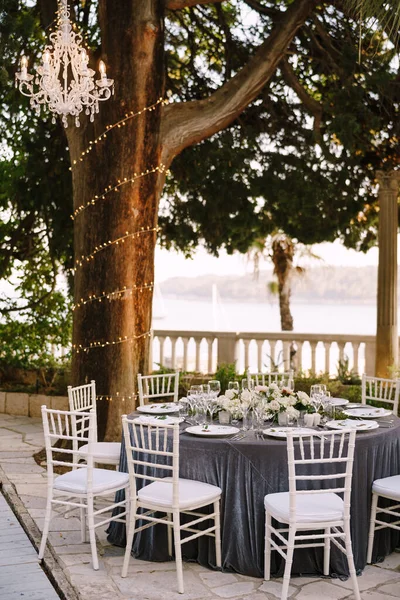 Recepción de mesa de cena de boda. Mesas redondas de banquete para invitados con mantel gris. Arreglo floral de rosas en el centro de la mesa. Guirnalda en troncos de árbol, candelabros de cristal cuelgan en el aire — Foto de Stock