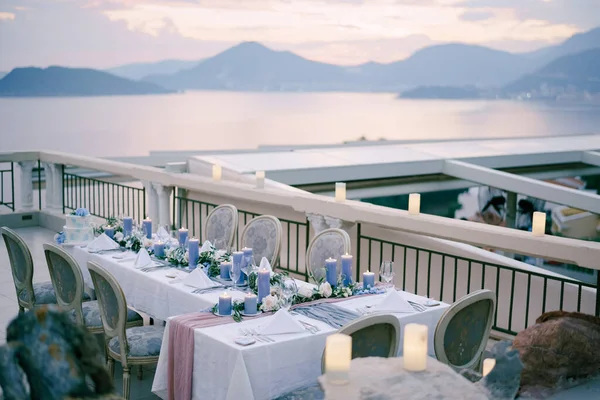 Recepción de mesa de cena de boda con muchas velas azules gruesas contra el cielo del atardecer sobre las montañas y el mar en Budva, Montenegro. Viejas sillas blandas en la terraza de verano de los restaurantes, al aire libre . — Foto de Stock