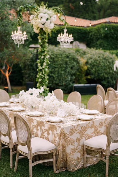 Weselne przyjęcie przy stole. Eleganckie stoły dla gości z kremowymi obrusami z wzorami, na zielonym trawniku, z wiszącymi nad nimi girlandami i żyrandolami. Krzesła z okrągłym tyłem — Zdjęcie stockowe