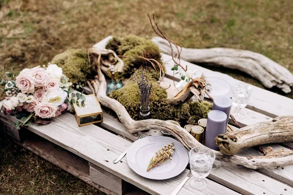 Resepsi meja makan malam pernikahan. Sebuah meja dadakan untuk dua palet konstruksi di rumput abu-abu di Islandia. Piring ungu terang, snags, lumut, lilin ungu, lavender, karangan bunga. Tampilan atas — Stok Foto