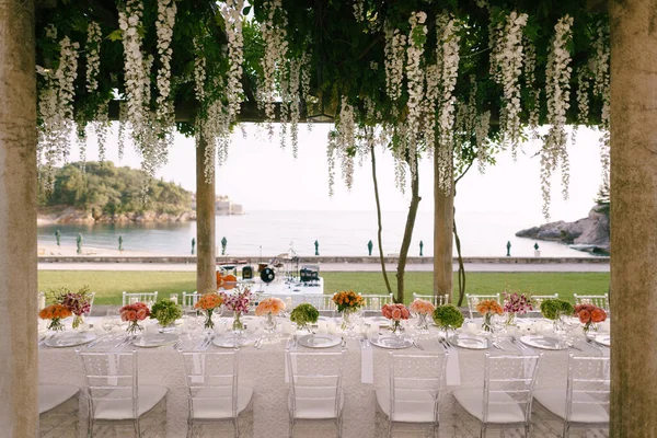 Recepción de mesa de cena de boda. Una mesa muy larga para invitados con un mantel blanco, arreglos florales, sillas transparentes de plástico de vidrio Chiavari. Bajo las viejas columnas con vides de glicina . — Foto de Stock