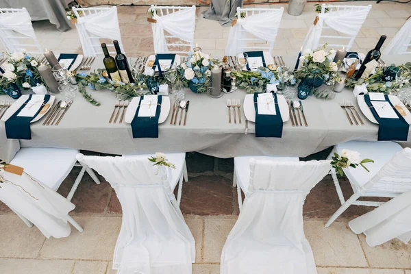 Recepción de mesa de cena de boda. Una mesa rectangular con un mantel gris, un arreglo floral con eucalipto, un plato blanco y una servilleta azul, sillas Chiavari decoradas con tela blanca y flores . — Foto de Stock